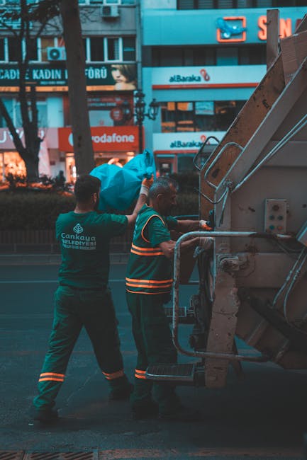 Two waste collection workers are positioned on a city street during dusk, loading a large blue plastic bag filled with rubbish into a flatbed truck used for private waste disposal. The worker on the l