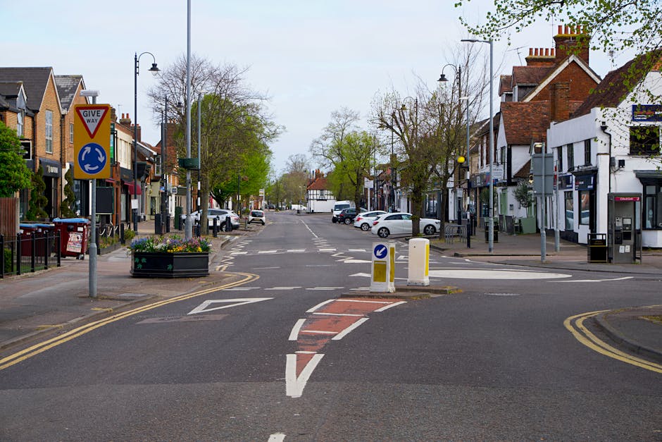A daytime street scene showing a quiet urban intersection with a central circular traffic roundabout, surrounded by residential and commercial buildings. The foreground features a road with painted wh