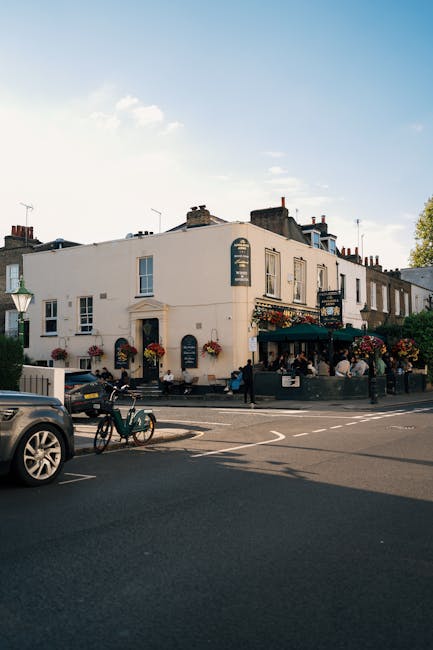 A street scene featuring a two-storey white building occupying the corner of a road, with large windows and a dark green awning covering an outdoor seating area, which is decorated with hanging basket